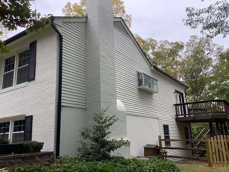 A two-story white house with a large chimney and a wooden deck surrounded by trees.